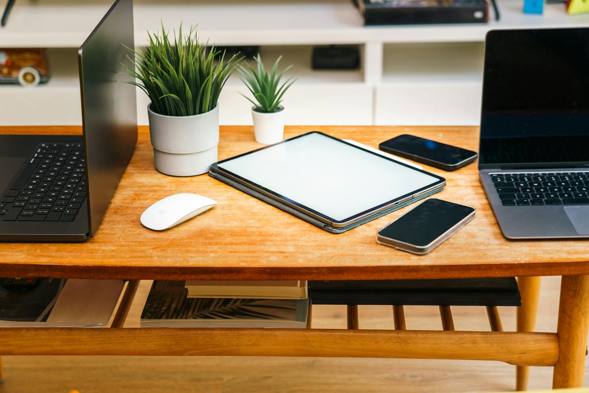 A stylish home office desk with laptops, tablet, smartphones, and potted plants.